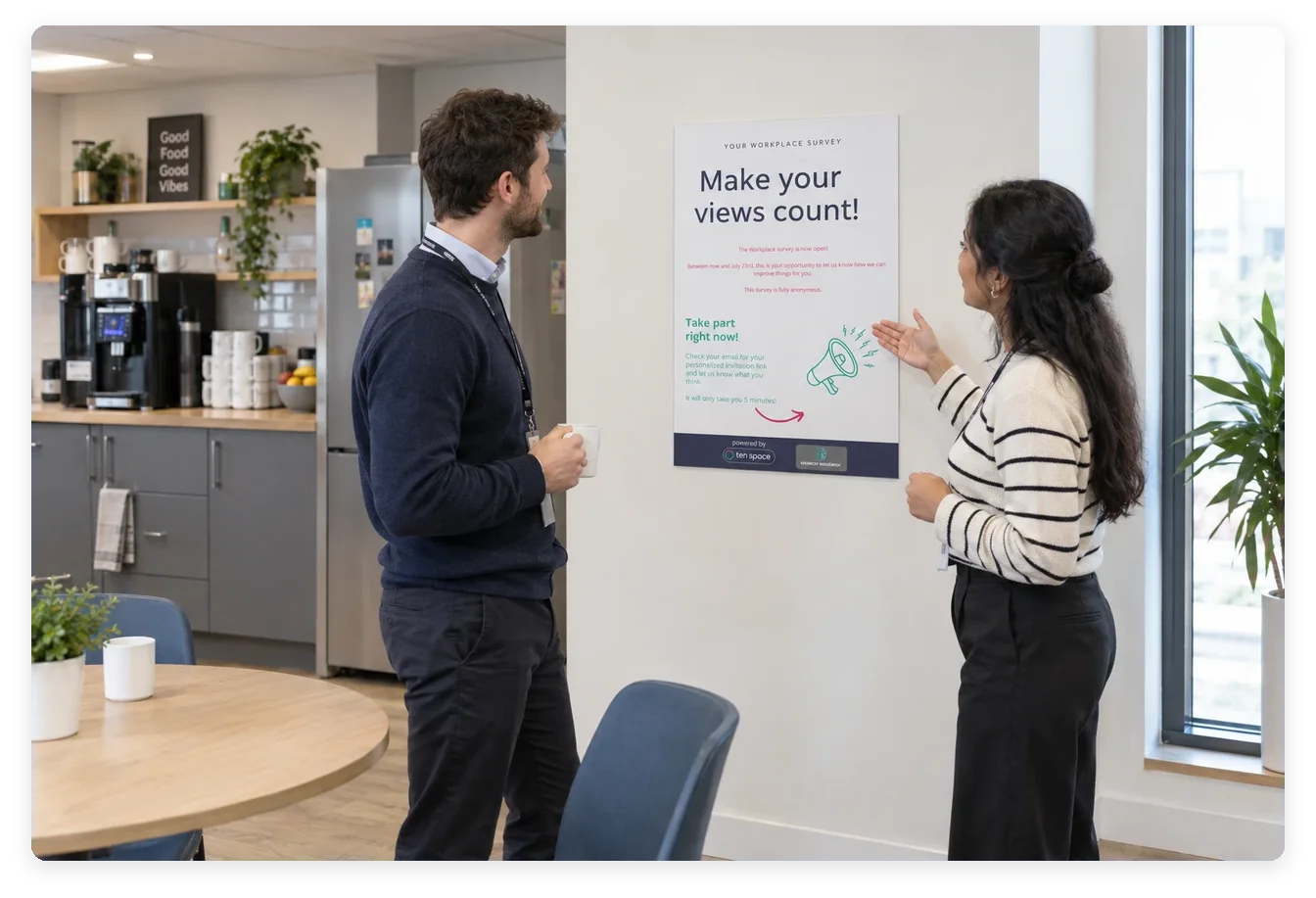 Two colleagues reading a workplace survey poster on a staff kitchen wall